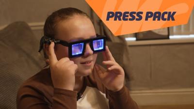 A girl wears a pair of glasses with thick black frames and lenses that are lit up with blue lights. She's pressing a button on the side of the glasses.