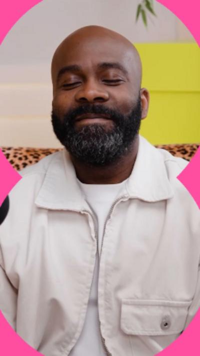 A man wearing a white T-shirt and a white denim jacket sits on a leopard-print couch. He has his eyes closed and is smiling. He has a full beard.