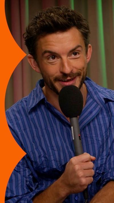 A man, Jonathan Bailey, wears a blue striped shirt and holds a microphone. He is smiling and looking at someone off camera.