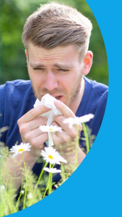 A man wearing a blue t-shirt sits in a field surrounded by daisies and sneezes into a tissue.