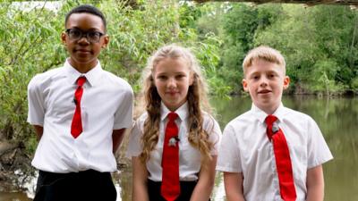 Three children in school uniform stand side by side facing the camera and smiling. Behind them is the bank of the River Wye and lots of trees