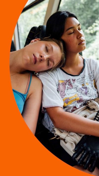 Two young women on a train, both sleeping, one with her head on her friend's shoulder.