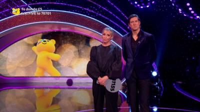 Mel Giedroyc and Vernon Kay stand on stage, both looking at the camera and dressed in dark clothes, with a cartoon Pudsey the Bear on a screen behind them