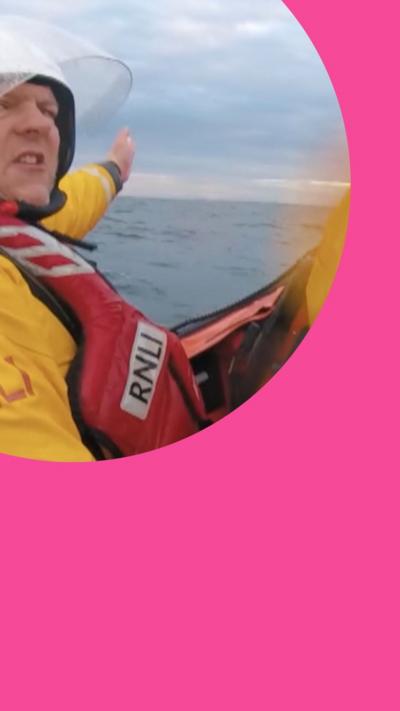 Two RNLI rescuers - dressed in lifejackets and yellow uniforms - are on a lifeboat in the sea. One points ahead