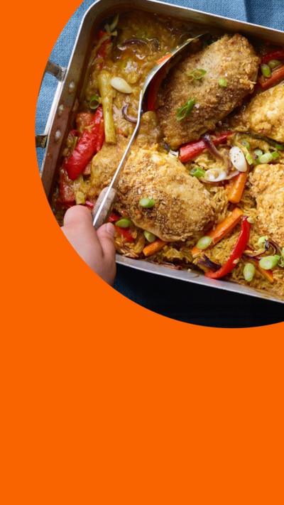 Four chicken breasts in a katsu curry sauce with chopped vegetables, all in a metal tray. A hand holds a big spoon