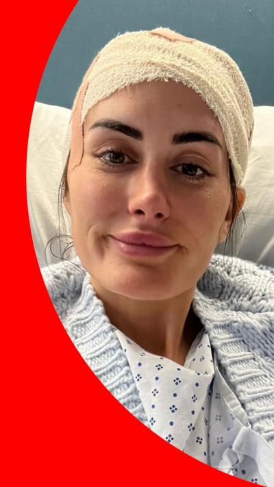 A woman with dark hair and eyebrows sit in a hospital bed. Bandages are wrapped around her head. She is wearing a hospital gown and a pale blue cardigan.