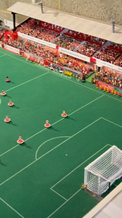 A replica model of Cheltenham Town Stadium with football players standing in the middle of the pitch and the stands full of fans.