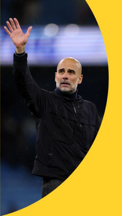 Pep Guardiola is waving. His right hand in the air and he is wearing a black coat.