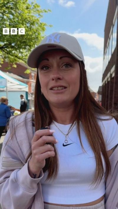 A woman with long blonde hair talking into a microphone. She has a baseball cap and tracksuit with a white t-shirt