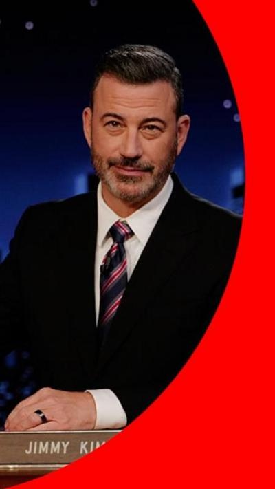 Jimmy Kimmel in a suit, sat behind a desk, on the set of his TV show