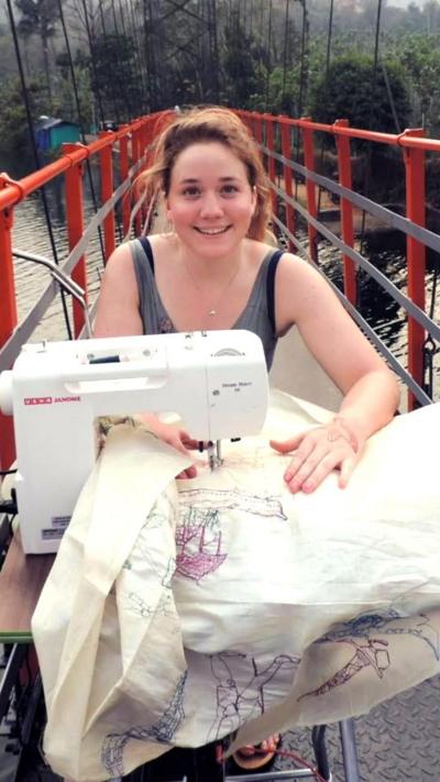 Harriet Riddell sits with her sewing machien and a large piece of fabric she is working on. She is sat outside on a bridge with red railings either side.