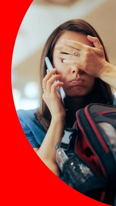 A woman with her hand over her face, talking on the phone while holding a bag in an airport lounge