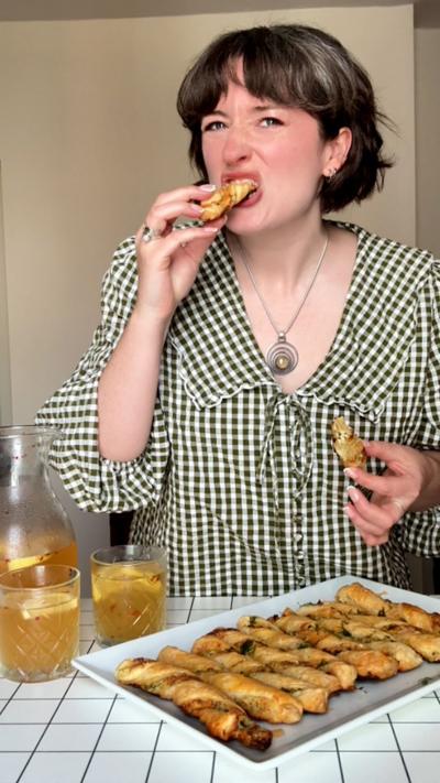 Charlie Thorne eating one of her cheesy pickle puff pastry twists while a big serving plate of them are on a table in front of her.