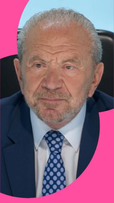 Alan Sugar looks at someone off camera. He has short grey hair and wears a blue suit.