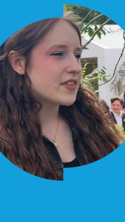 A young woman with long brown hair looks to the side of the camera. There is a white tent in the background. 