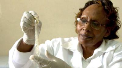 Image of a woman in a white lab coat, glasses and gloves holds a condom