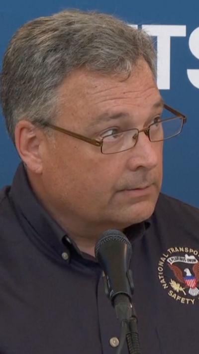 A man wearing glasses and a blue button up looks to his left while sitting in front of a microphone.