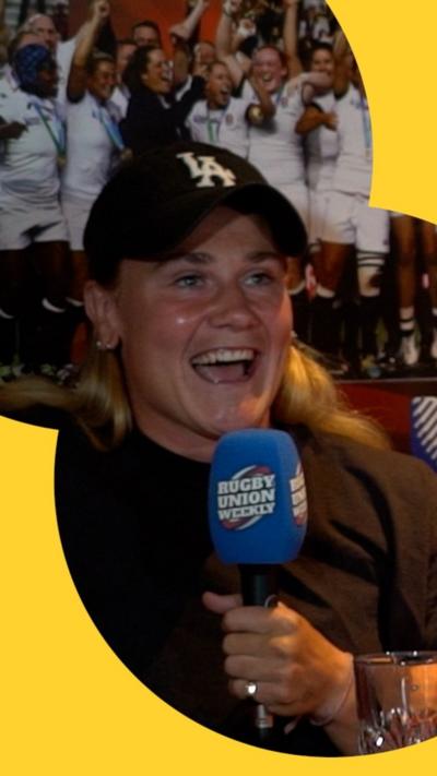 A women wearing a black cap holds microphones while sitting on a red sofa.
