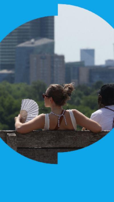 A photo of the back of a woman's head. She's sat on a bench holding a hand fan, looking out at a city.