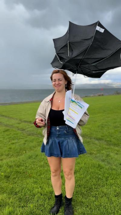 A girl with auburn hair is wearing a cream jacket with red tartan lining, a black top, a denim skirt and is holding a broken black umbrella while standing in front of the Clyde in Port Glasgow.