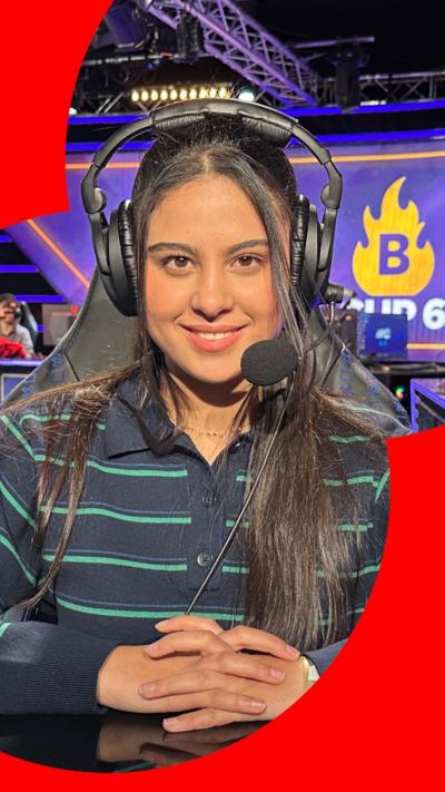 A young woman is sitting in a computer chair, in an e-sport venue. She is looking directly into the camera, smiling. She is wearing a striped shirt and a headset.