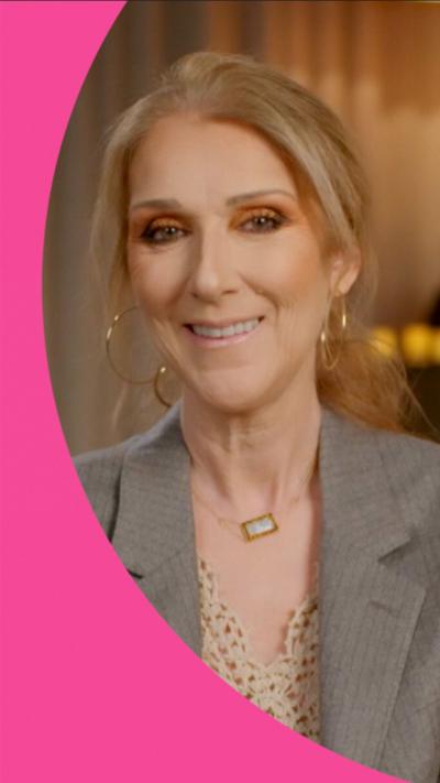 Céline Dion sitting on a chair with flowers behind her, smiling and wearing a grey blazer with a cream top