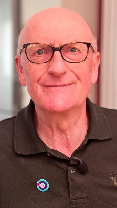 A bald man looks into the camera. He is wearing black rimmed glasses and is wearing a brown polo shirt.