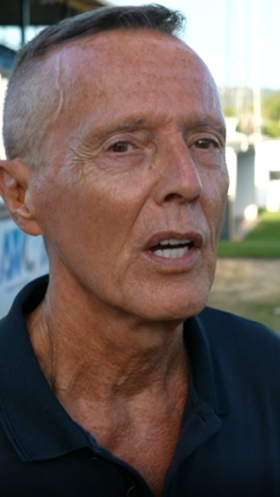 A tanned man wearing a dark polo top is talking to a camera on a football pitch.
