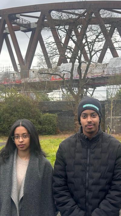 Two people - a woman and a man - stood in front of a HS2 viaduct. The woman has black hair and a grey coat. The man has a black hat and black coat. Behind them is a patch of grass then a large rust coloured structure