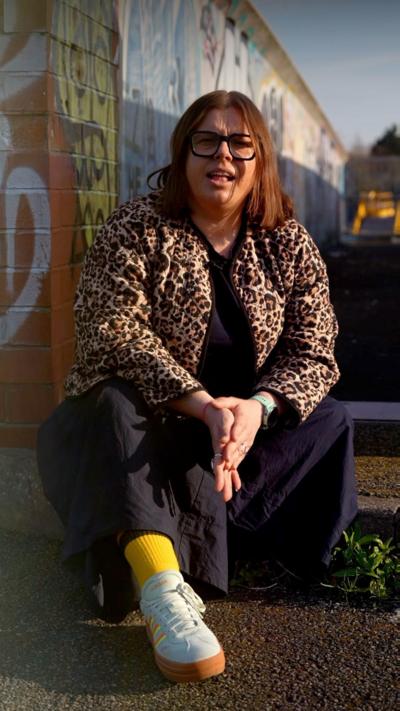 A woman with spectacles wears a patterned jacket and sits on a step in front of a grafitti-covered wall