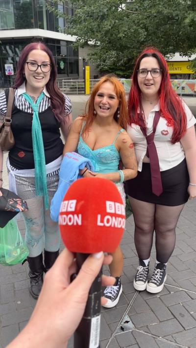 Three young women stand excited in front of a BBC London branded microphone outside Wembley Stadium.