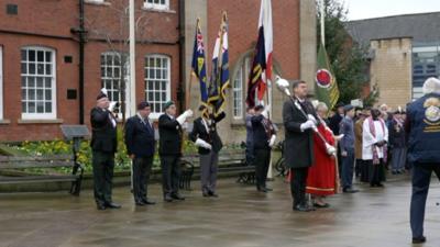 People in military dress holding flags and a sceptre outside a red bricked building in Wrexham