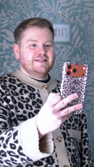 A man in a leopard print cardigan holding up a phone in a leopard print case