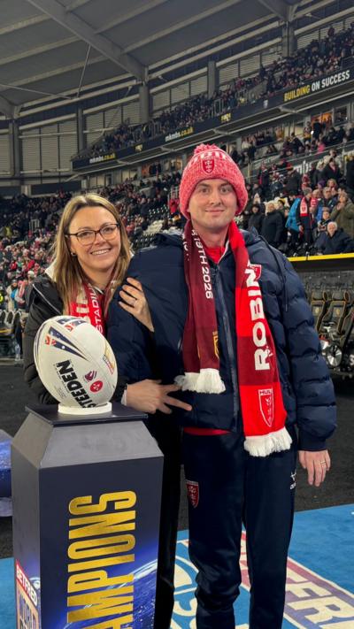 Craig Eskrett and his wife standing beside a display plinth holding a rugby ball inside a large, crowded stadium. They are both wearing warm clothing and team scarves, with spectators filling the stands behind them.