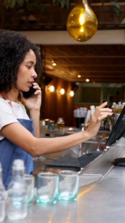 A woman wearing an apron behind a bar holds a phone to her ear with one hand and is typing on a screen with her other