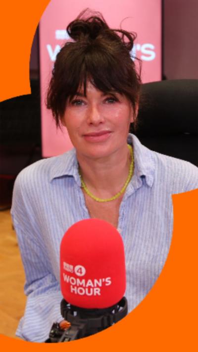 A woman in a brown-haired bun and linen blue shirt sits in front of a microphone.