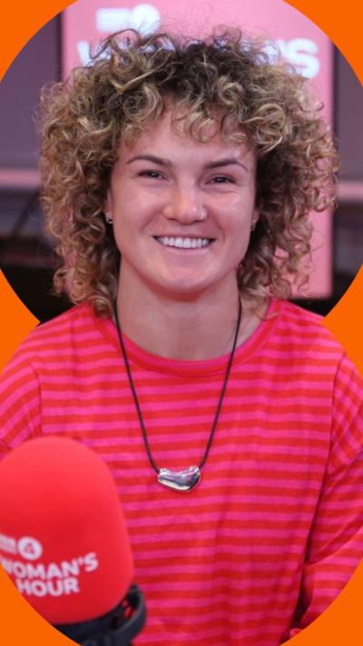 Ellie Kildunne smiles at the camera. She is wearing a red striped, long-sleeve top. She sits in-front of a red BBC Woman's Hour microphone.