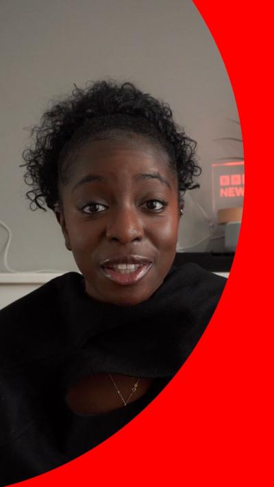 A woman with curly black hair wearing a black top talks to the camera.