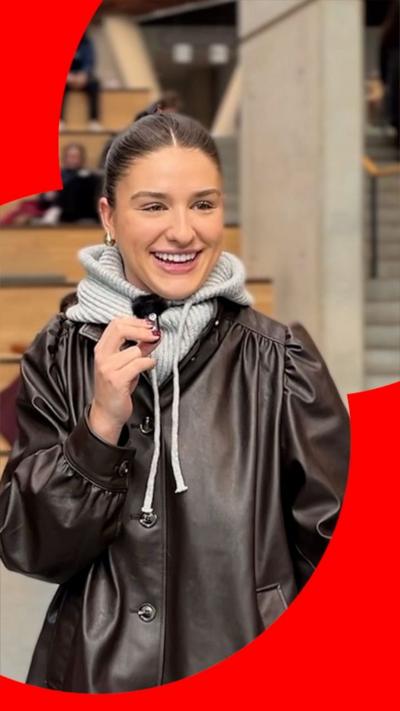 Student in a brown leather jacket with a grey hoody underneath. She has brown hair tied back and is holding a small microphone.