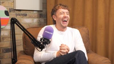Roman Kemp sat on chair in front of a purple microphone and laughing