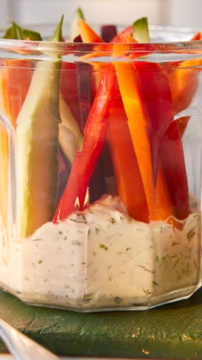 A jar with creamy dip at the bottom with veg sticks at the top on a green chopping board.