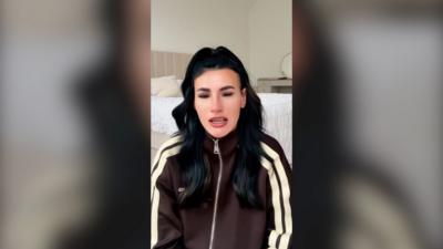 A woman, Brittany Miller, with dark hair and a brown sports jacket, looking tearful to camera. A still from a video.