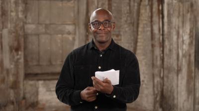 Paterson Joseph, a black man with a bald head, smiles at the camera. He is wearing black glasses and a black shirt. He is holding a piece of paper in his hand