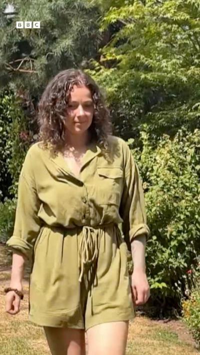 A woman in a green outfit walking in a garden. She has long brown, curly hair