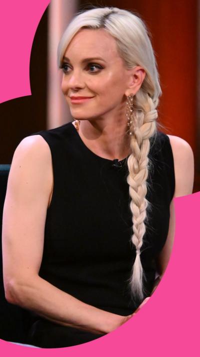 Anna Faris smiles at someone off-camera, she has long blonde hair in a side plait. She wears a sleeveless black top.