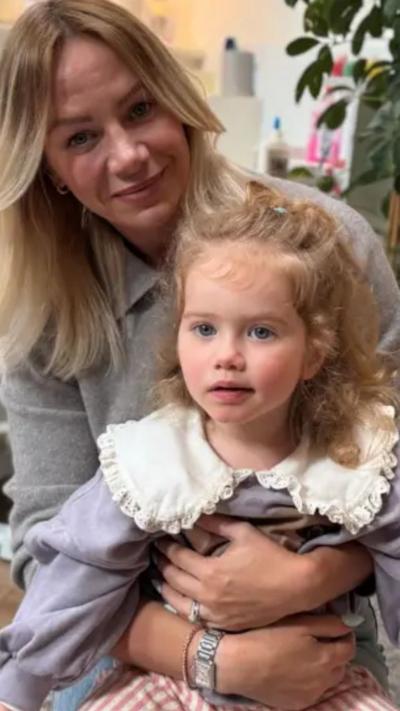 A young girl in a lilac top with white collar being cuddled by her mum, who has long blonde hair and is wearing a grey top