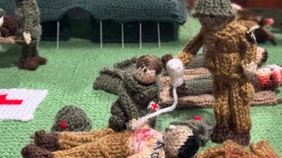 A knitted medical facility depicting an injured soldier receiving treatment during the D-Day landings at Utah Beach. A knitted topless and bleeding soldier is receiving treatment from a medic wearing a green knitted uniform. Another soldier wearing a light brown uniform is standing over the pair holding an IV drip. Knitted injured and wounded soldiers are also lying on the green knitted base, while another injured soldier is being carried on a stretcher beside a dark green medical tent.