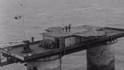 A view of Sealand from a helicopter, which is a platform on top of two massive concrete legs.. Bob Wellings and Roy and Joan Bates are waving from the roof.