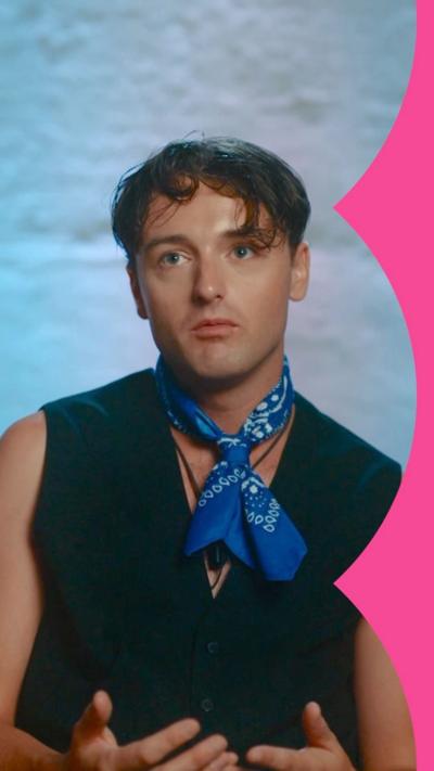 Adam looking serious, he is wearing a black vest and a blue necktie