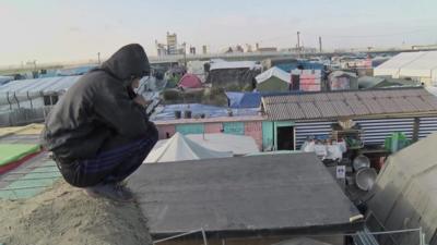 A person perches above the roofs of the camp.